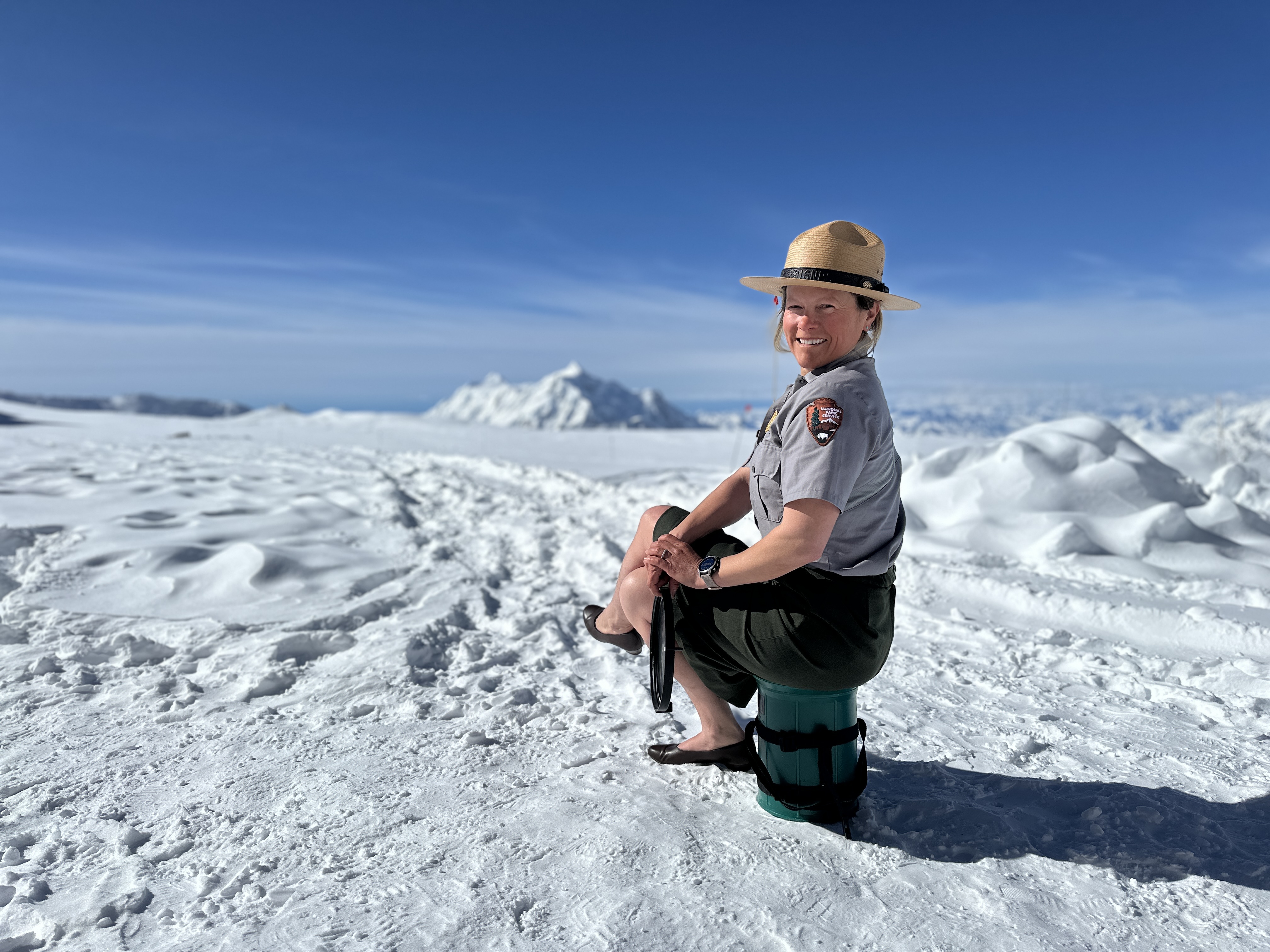 A smiling park ranger in an NPS flat hat, skirt, and pumps sits atop a green Clean Mountan Can, with a snow covered peak in the distance