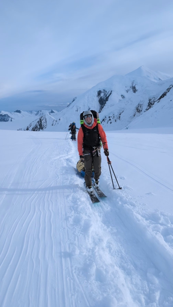 Two rangers skiing toward the camera. The ranger in front is smiling and wearing an orange jacket and black snow pants.