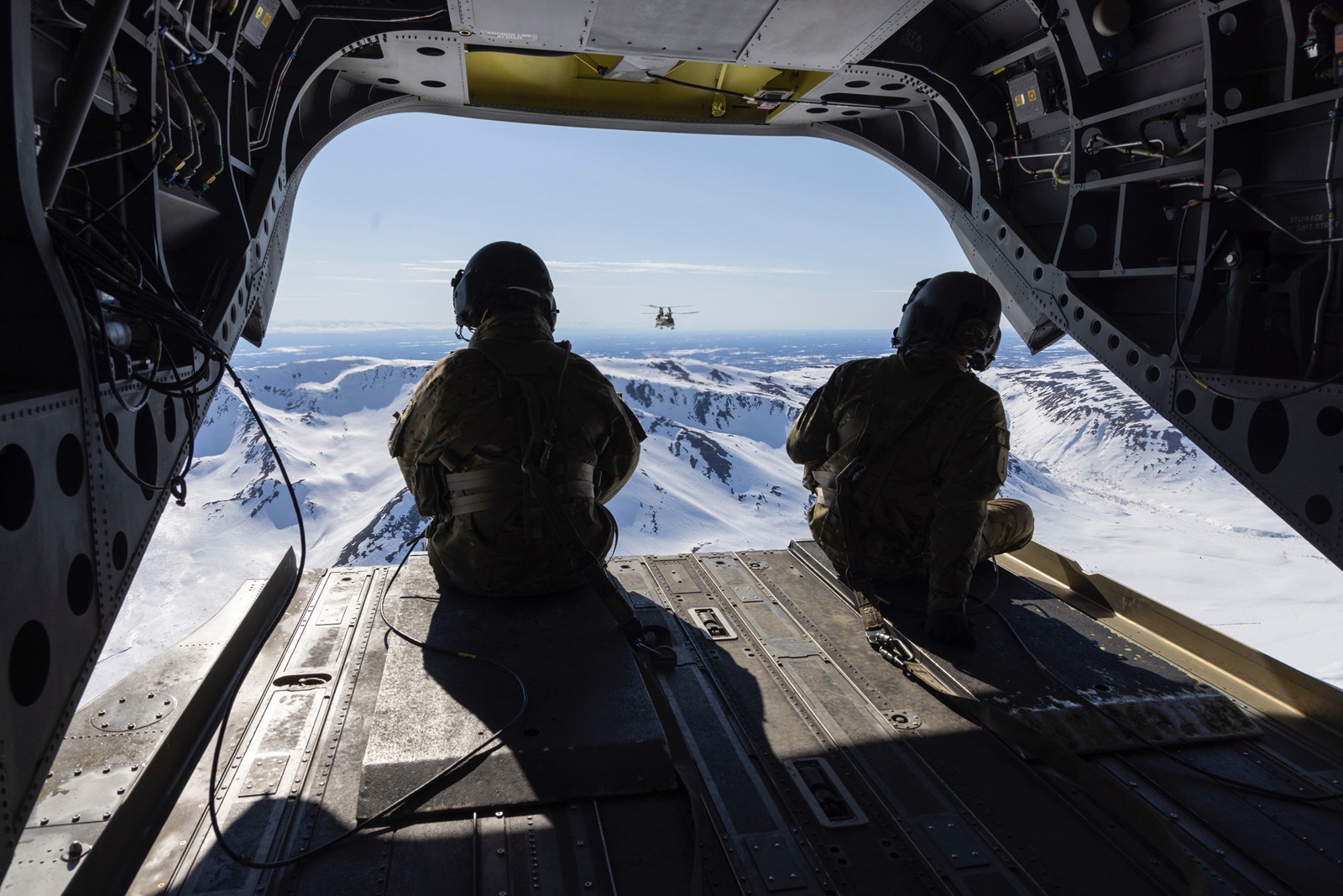 Two soldiers sit on the open back gate of a Chinook helicopter as it flies into the mountains