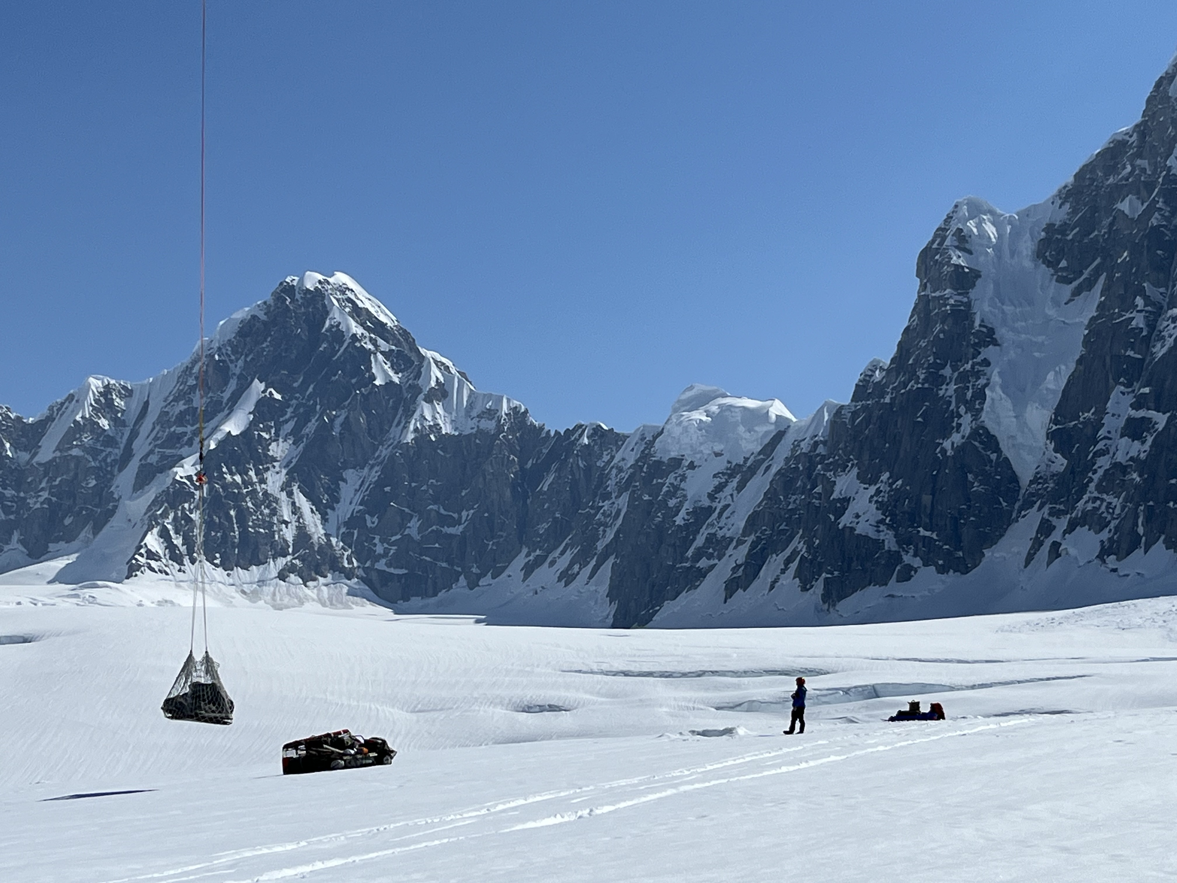 A silhouette of a ranger standing on a glacier observing a net load of equipment suspended at the end of a long rope line.