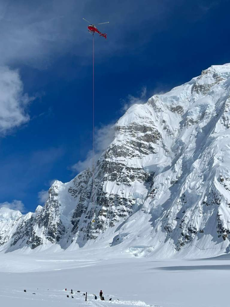 A red helicopter hovers high above a snowy landscape with a rope lowered to a ranger standing below it in front of a large mountain peak and blue sky.