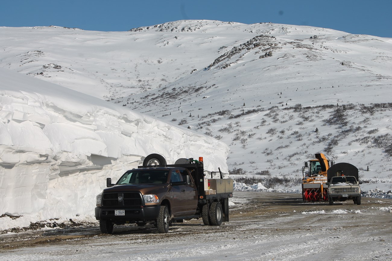trucks on a dirt road near a hillside covered deep in snow