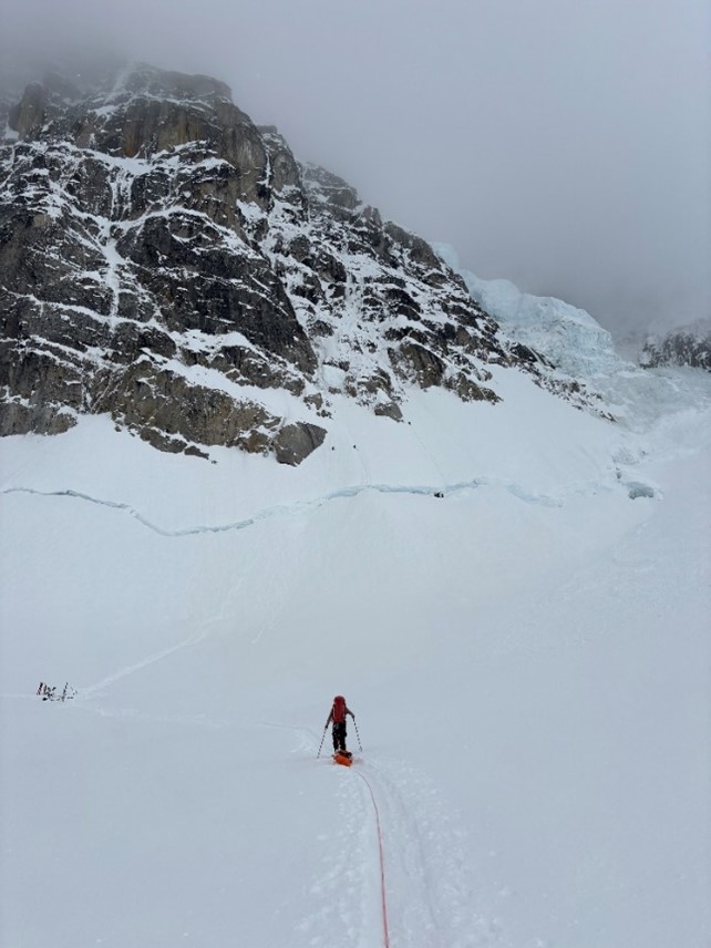 A ranger makes their way across a snowy landscape toward a grey mountain. 