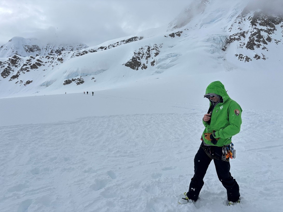 Ranger in a green jacket standing on a snowy landscape with other rangers approaching in the distance. 