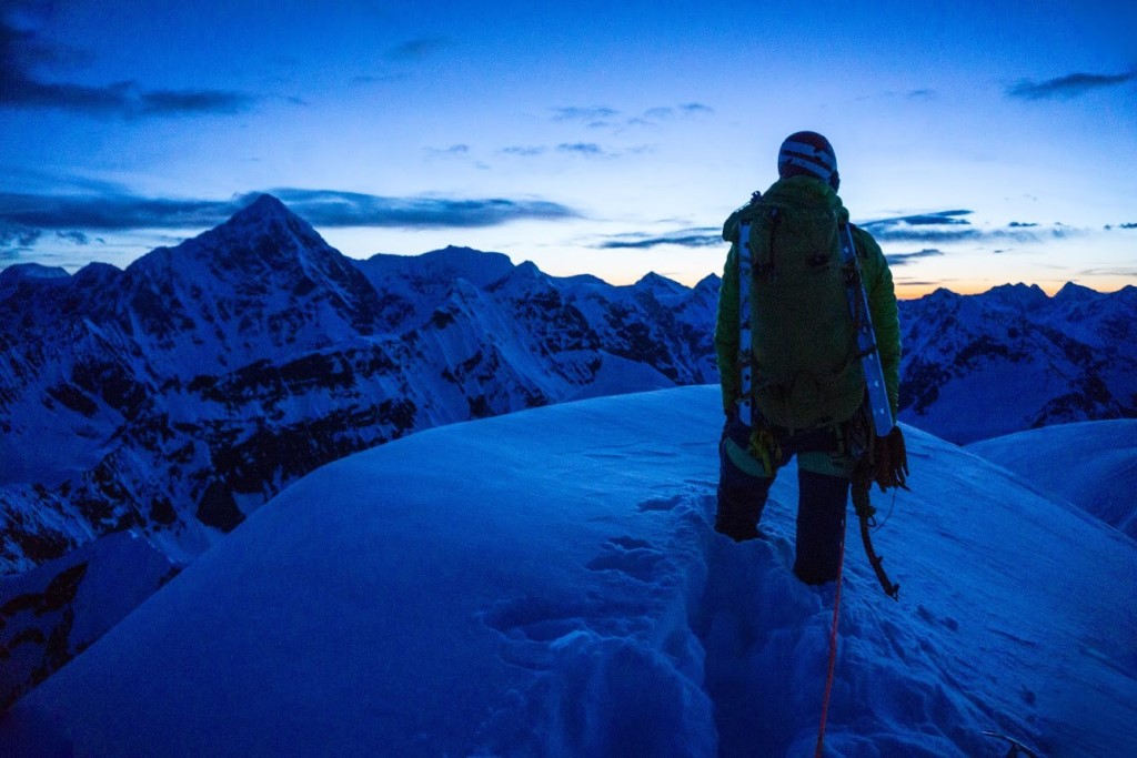 A climber on top of a snowy peak stares off into a distant sunset