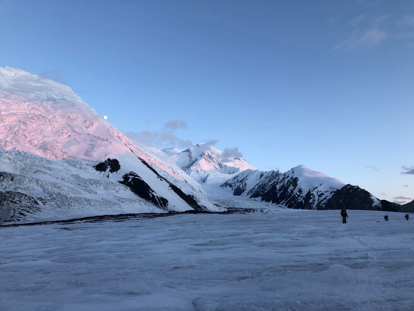 Three distant climbers move along a flat, smooth glacier surface, with pink alpenglow illuminating the surrounding snow covered ridge.