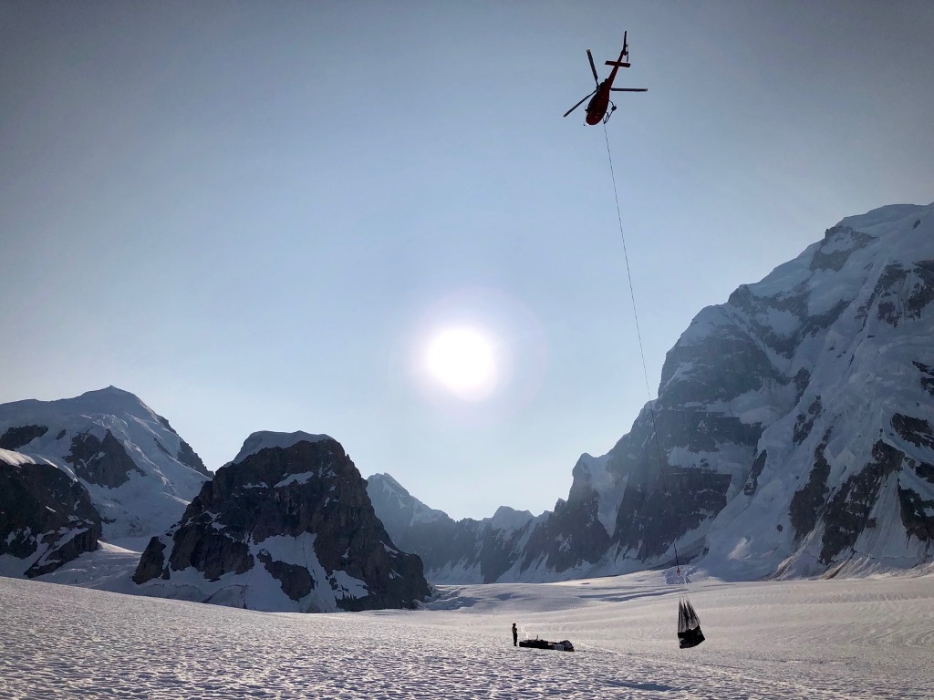 A helicopter hovers over a netload of gear suspended by a rope