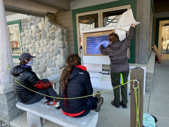 Two climbers seated on porch bench listen to a ranger describe mountain features on an outdoor display board 