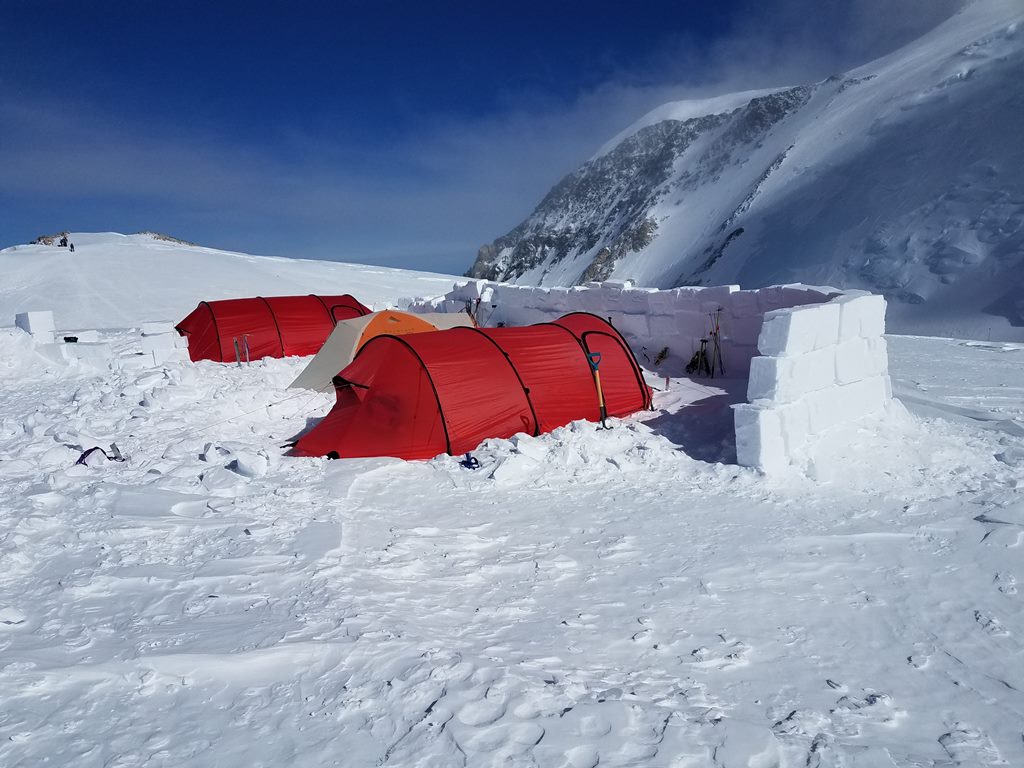 Three tents on a glacier shielded on one side by a snow wall