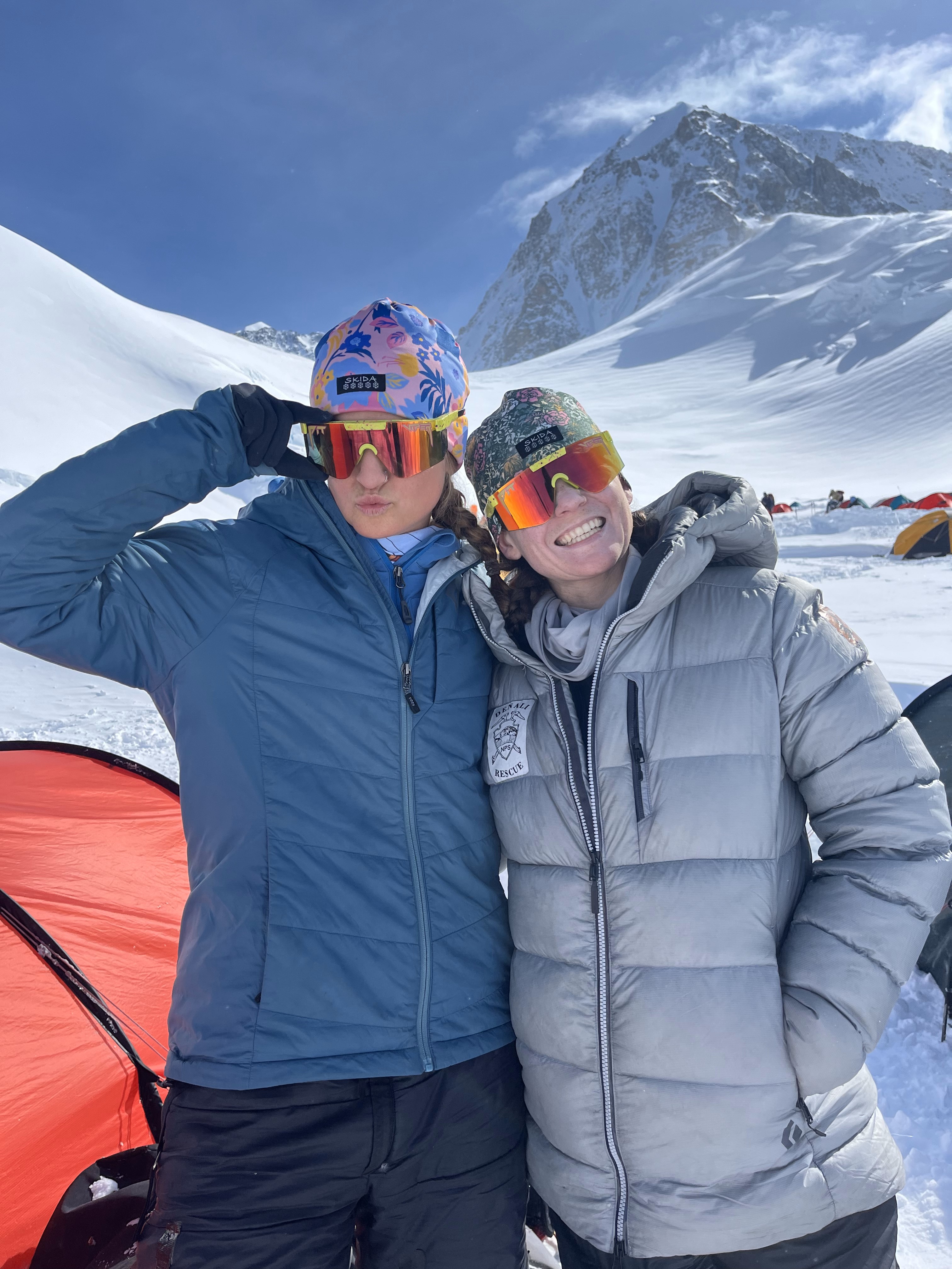 2 rangers wearing mirrored ski glasses and down coats pose in front of red tent and a snow covered mountain