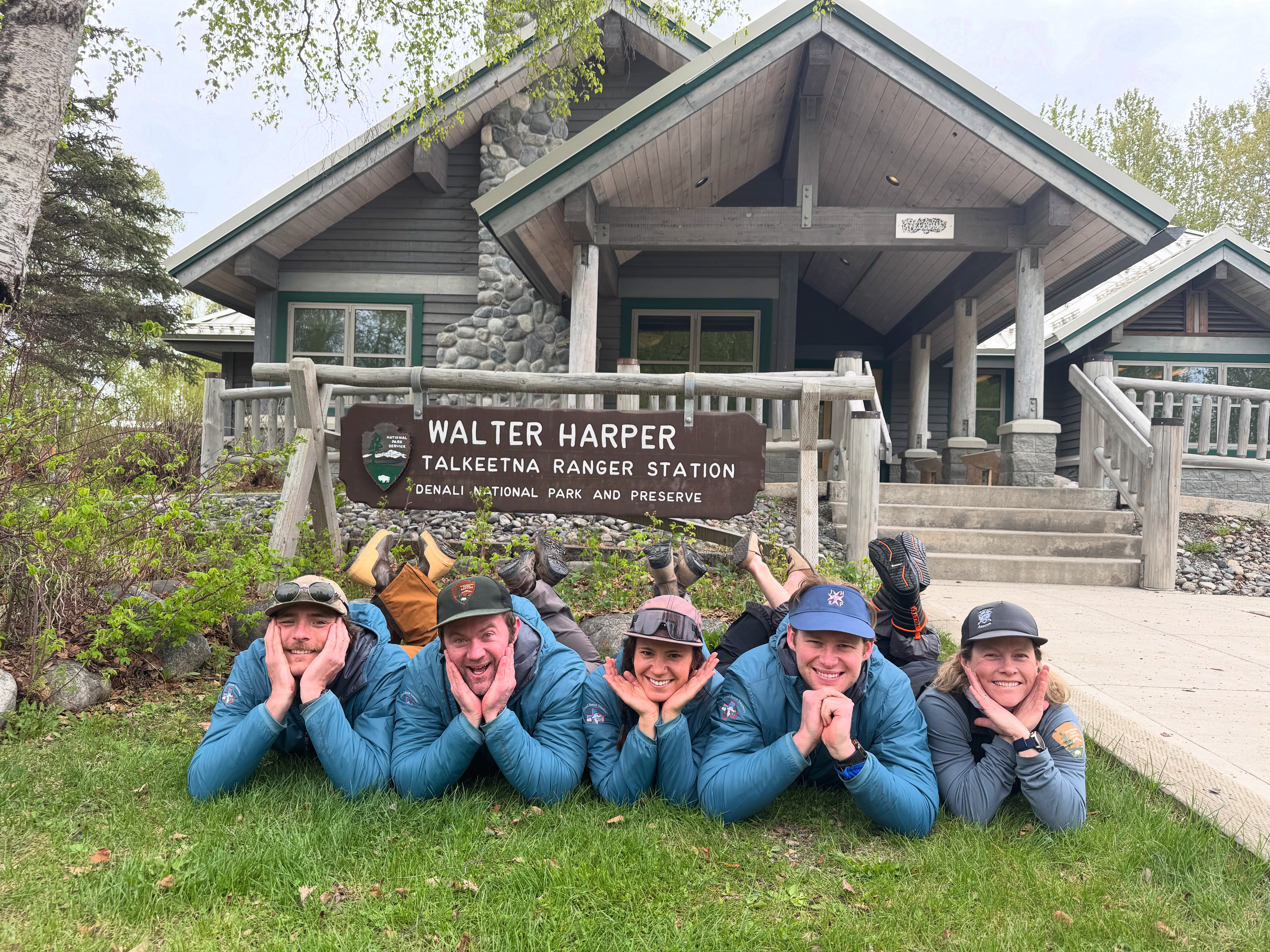 5 rangers lie on the ground with their chins in their hands in front of the Walter Harper Talkeetna Ranger Station