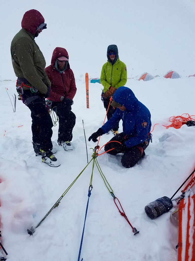 A ranger kneels in the snow explaining a rope rigging system to fellow rescuers