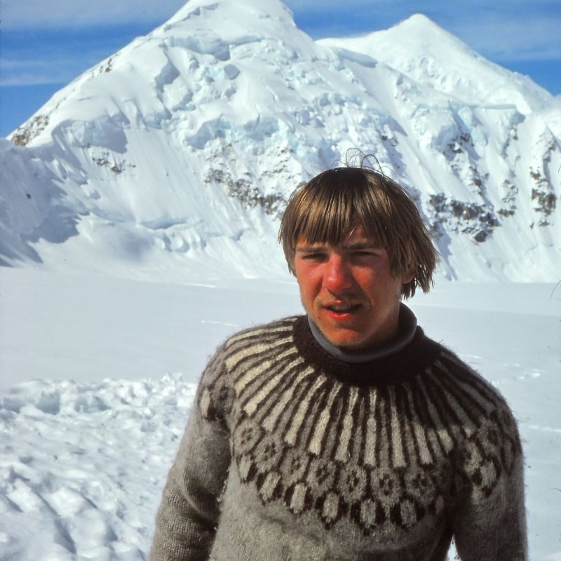 Man with sandy blonde hair wearing a brown fair isle sweater in front of a snowy mountain peak