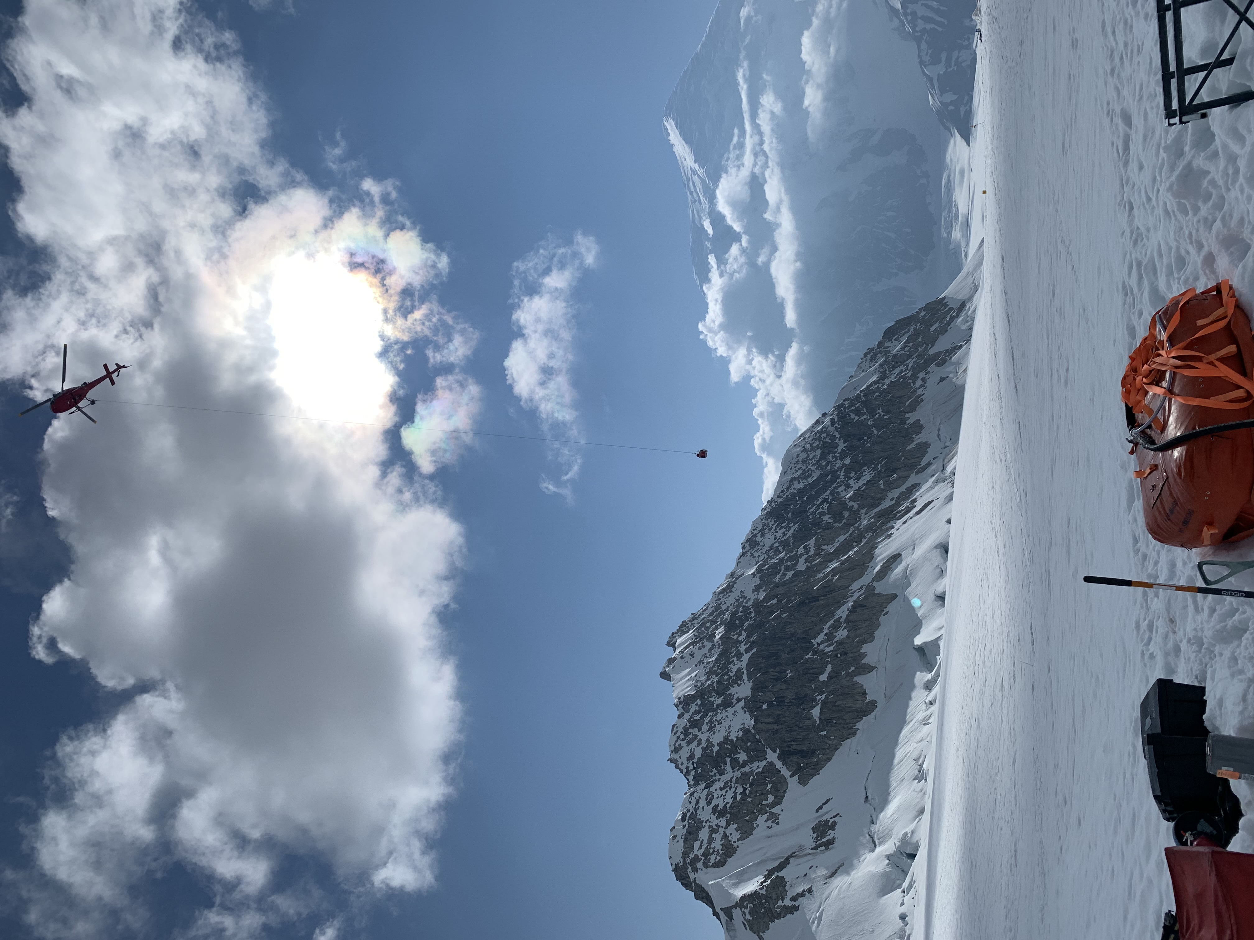 A helicopter over a glacier carrying a long rope line with a basket at the end of it 