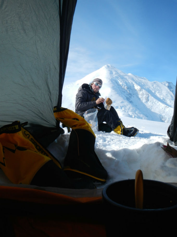 Looking out a tent to a snacking climber seated in the snow