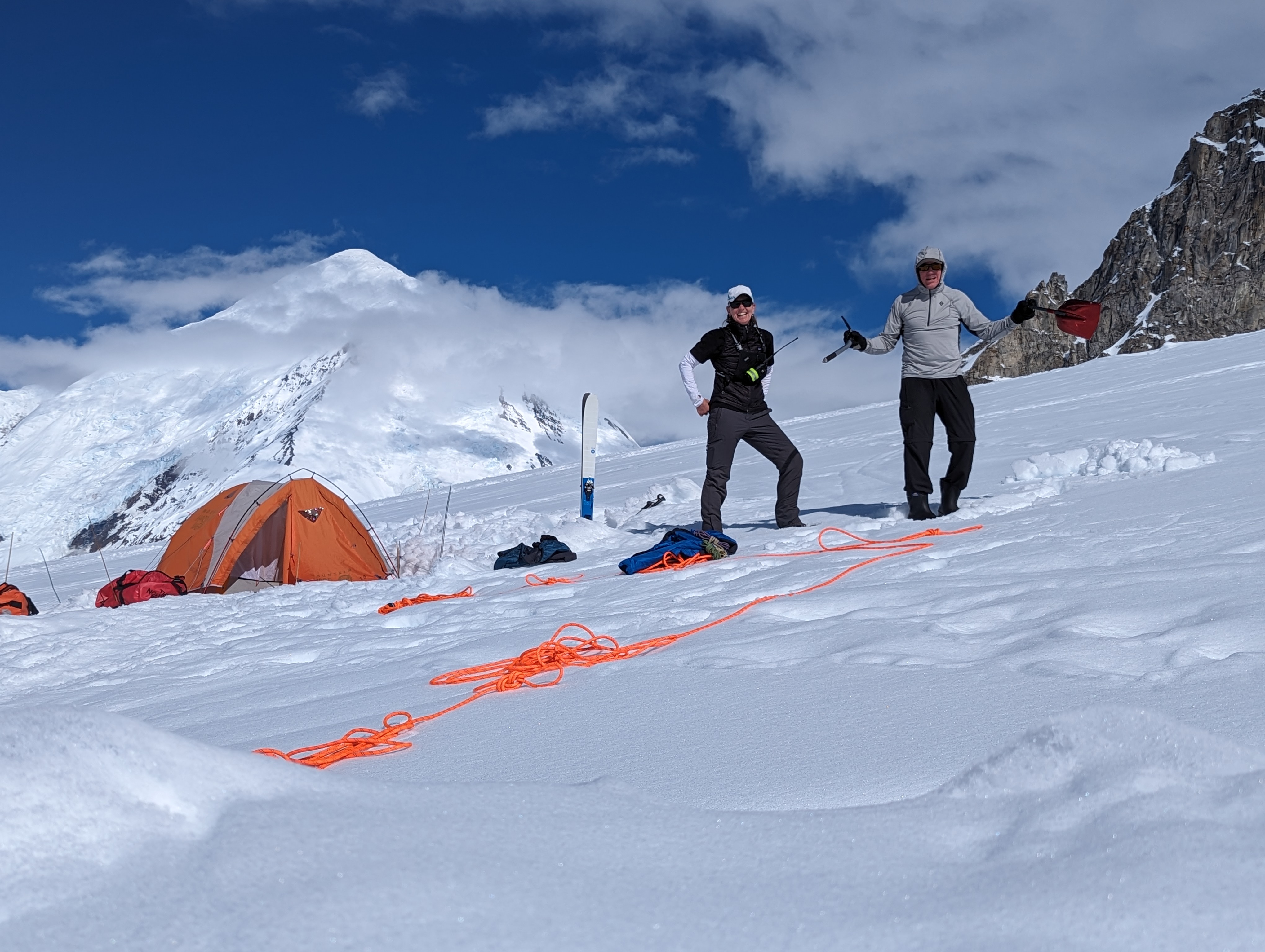 Two climbers stand on a glacier near a pile of ropes and a tent