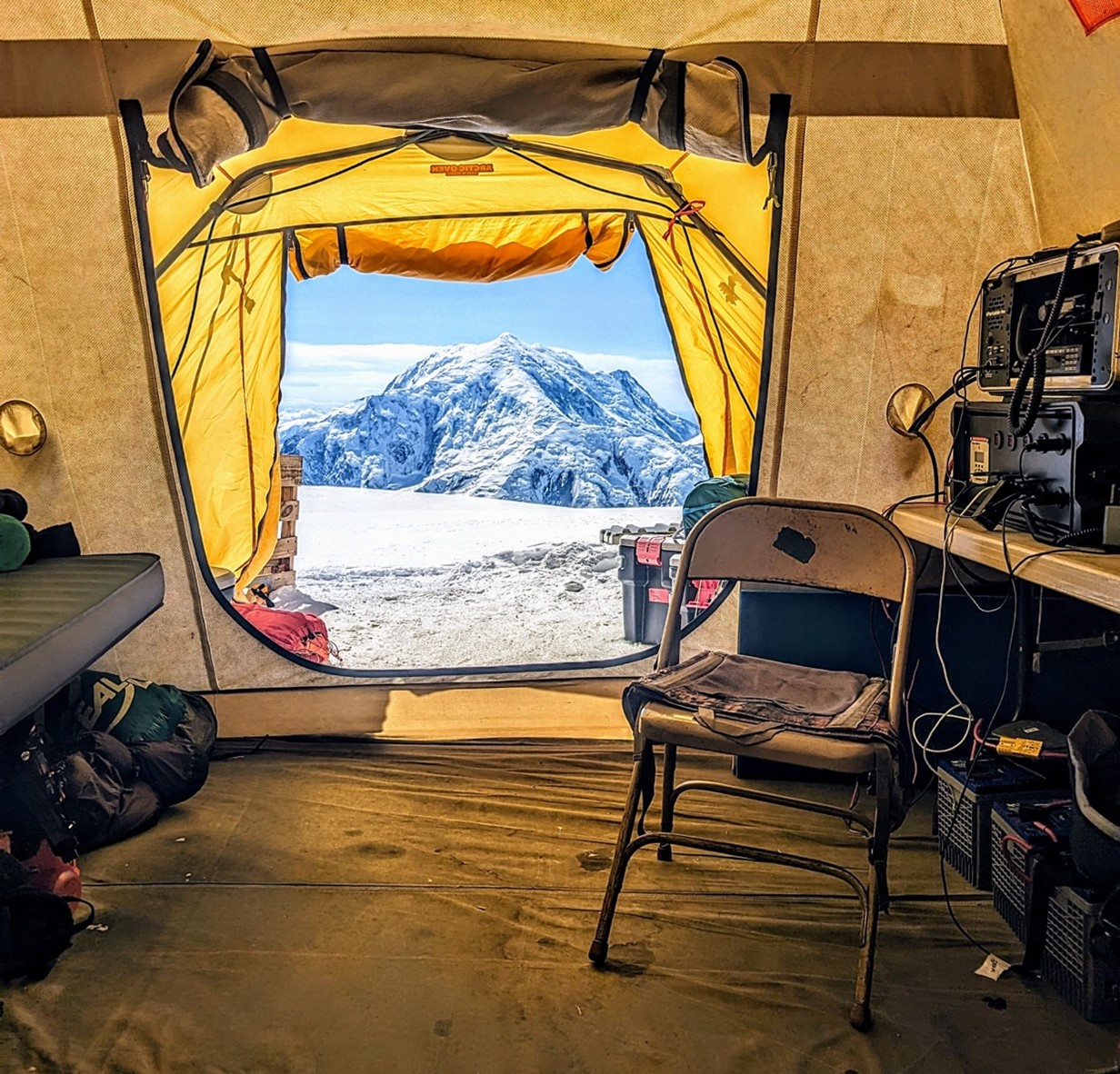 View looking out a yellow tent at a mountain in the distance. A metal folding chair and a radio console sits inside the tent.