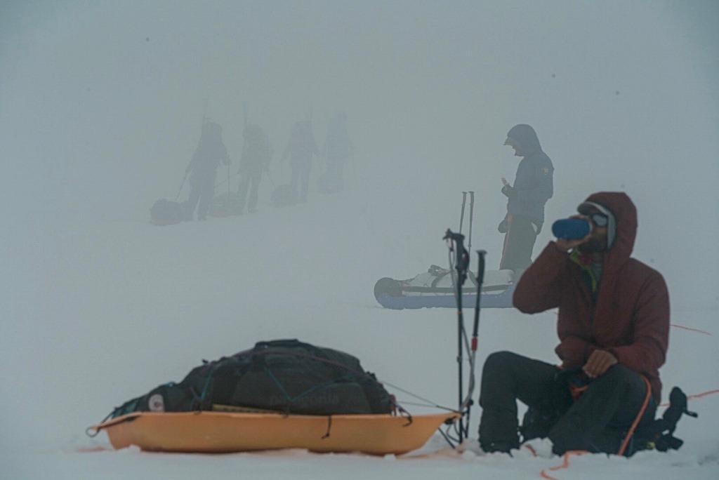 On a foggy glacier, a climber sits next to his sled and enjoys a bottle of water 