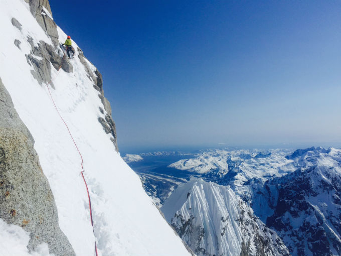 A roped climber ascends a steep rock and snow face with dramatic views of the Alaska Range in the distance