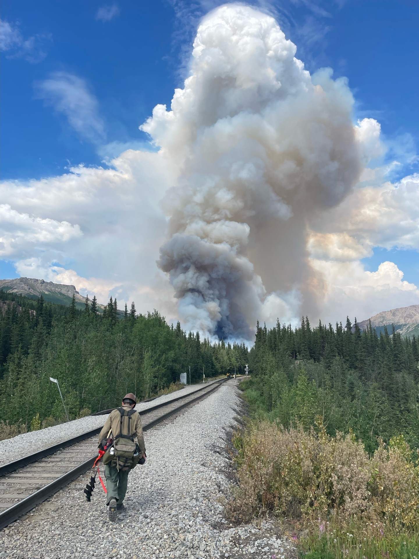 Wildland firefighter walks along railroad tracks toward a large plume of smoke