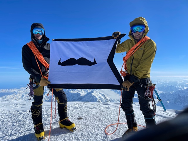 Two men stand on a mountain summit holding a flag with a mustache on it