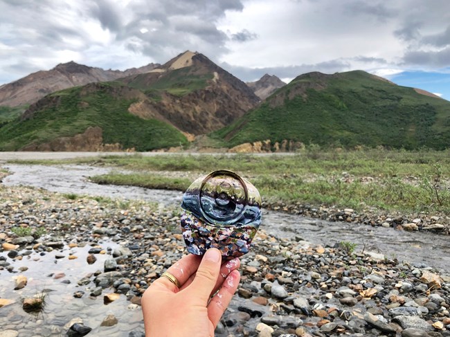 A hand holds out a smashed drink can that is painted to blend in with the green, brown mountains and rocky river in the landscape.