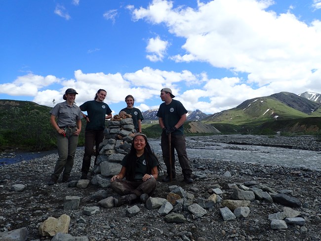 Four YCC participants and one ranger pose by a large rock cairn along a gravel river bar.