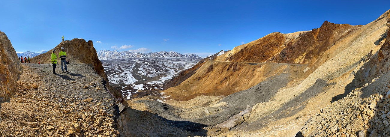 A panorama photo taken from the east edge of the Pretty Rocks landslide. Two workers in safety vests stand near the edge of the landslide and the mountain side drops away below them.