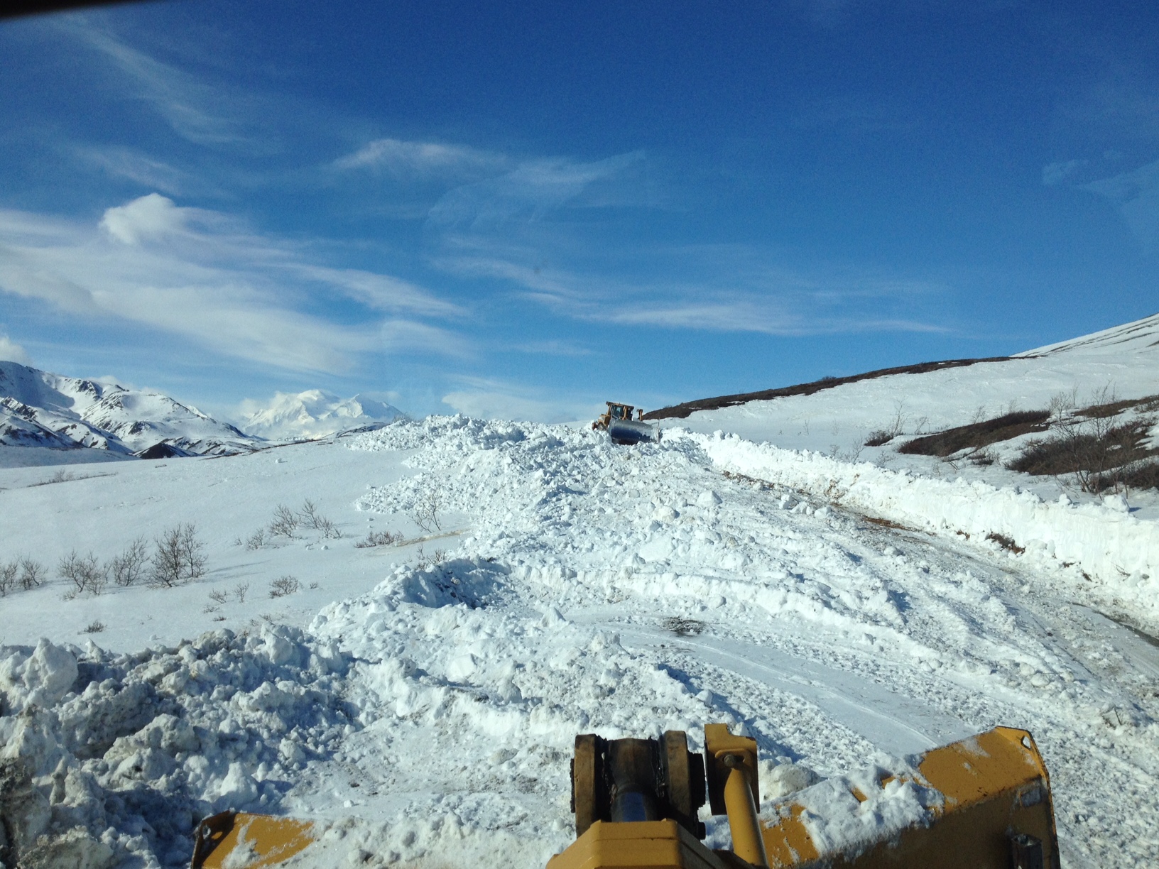 two large machines plow a snowy road on a hillside under a beautiful blue sky