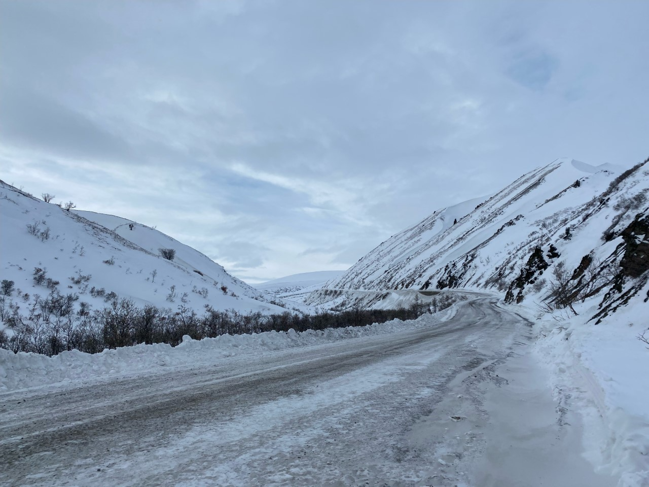 A road that has been mostly cleared of snow gradually climbs uphill at the base of a mountain. A thin layer of compacted snow remains on the road surface, but lots of gravel underneath can be seen through the snow.