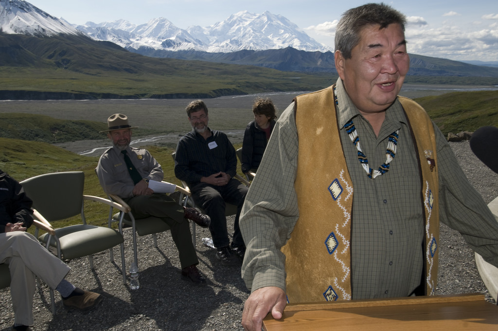 an alaska native man speaking at a lectern outside in front of several seated people. denali, a vast snowy mountain, is in the distance
