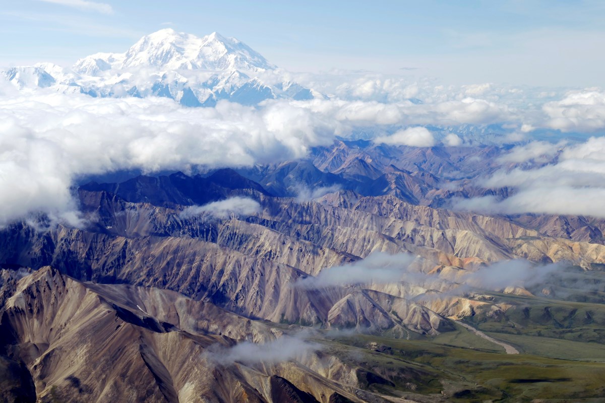 aerial view of clouds hanging over low mountains, with one huge snowy mountain towering above