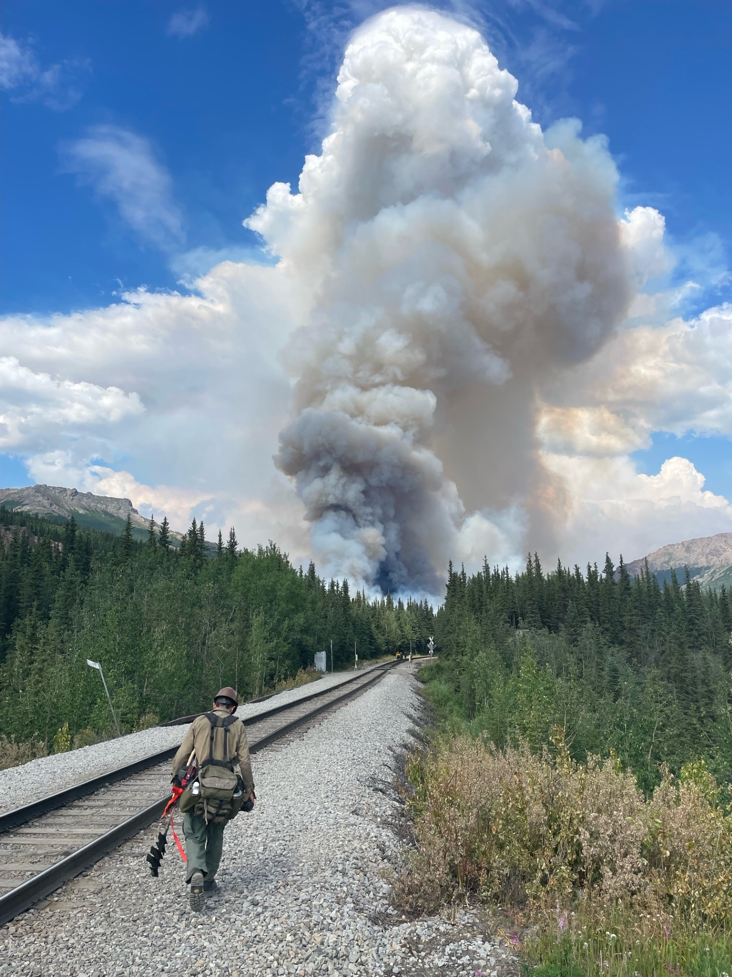 A firefighter walks along railroad tracks toward a large plume of smoke.