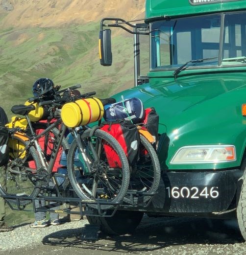 A bike rack mounted to the front of a green bus carrying two bikes. The bikes are loaded with camping gear.