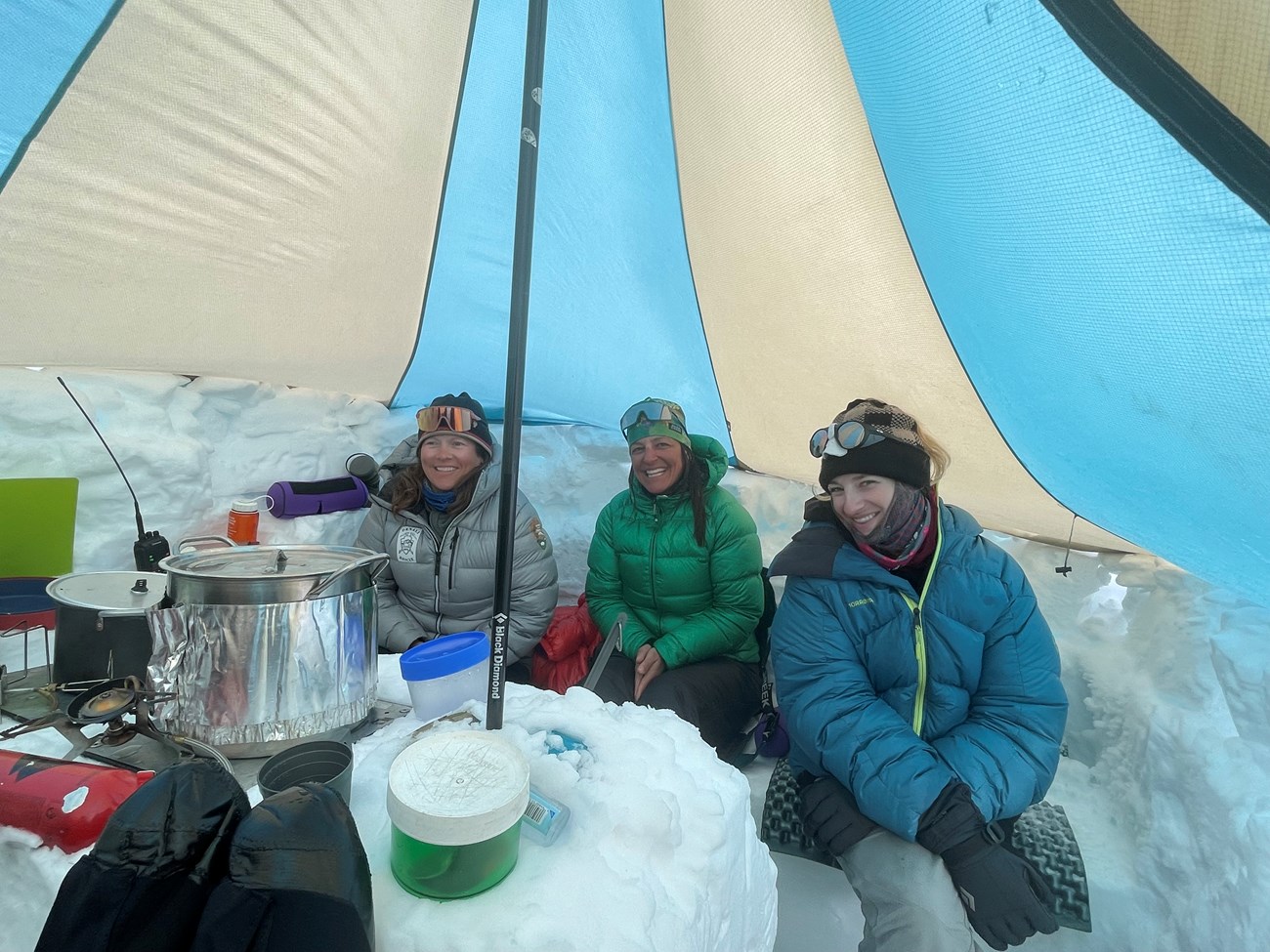 Three smiling climbers sit in a tent around a cookstove