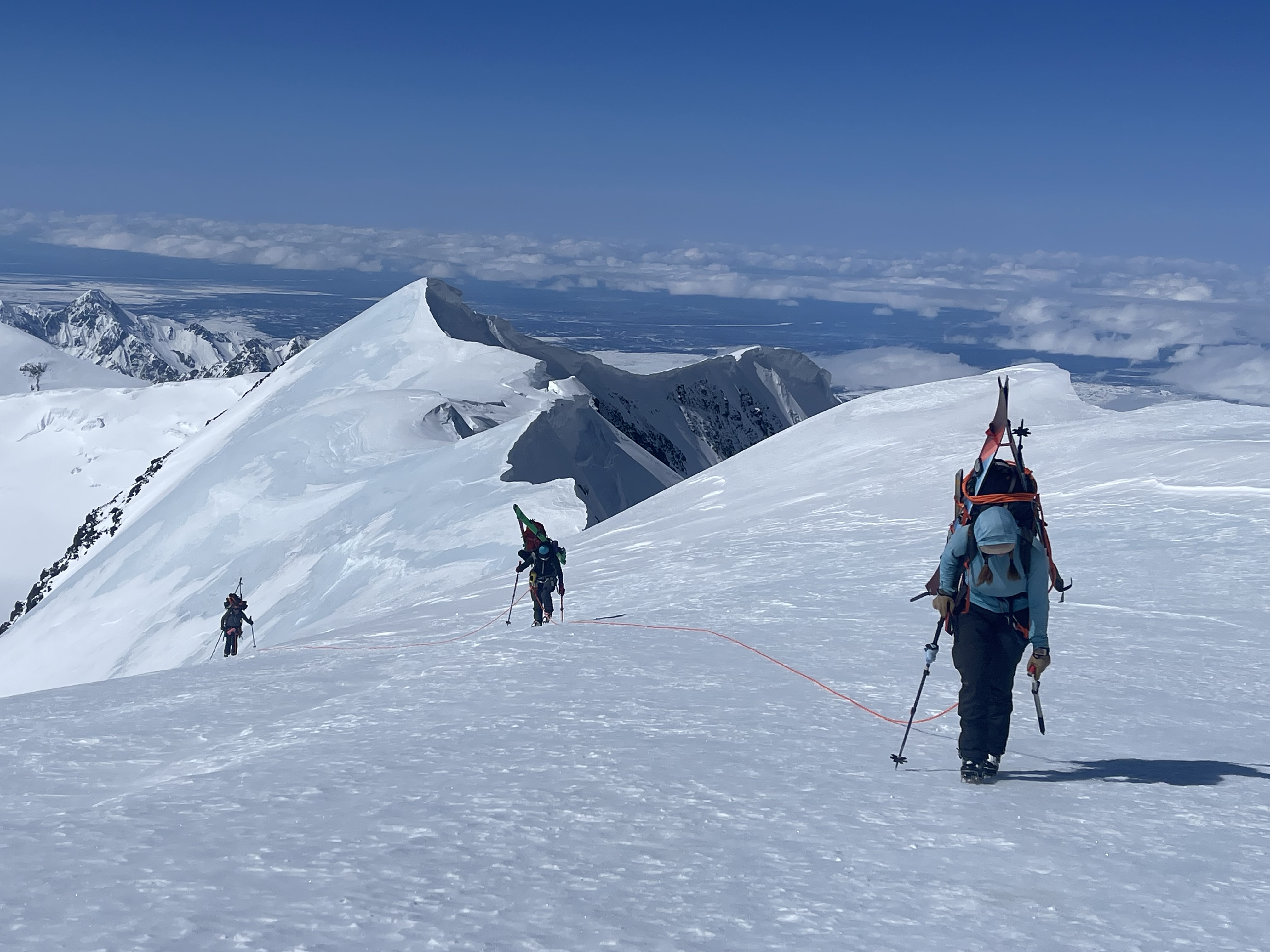 Three roped climbers move uphill with heavy backpacks with sweeping mountain views in the background