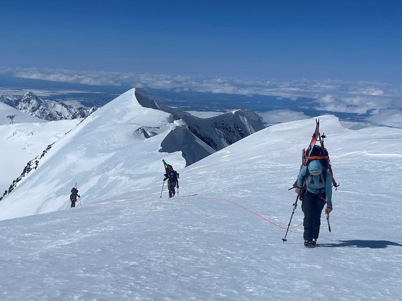 Three roped climbers move uphill with heavy backpacks with sweeping mountain views in the background
