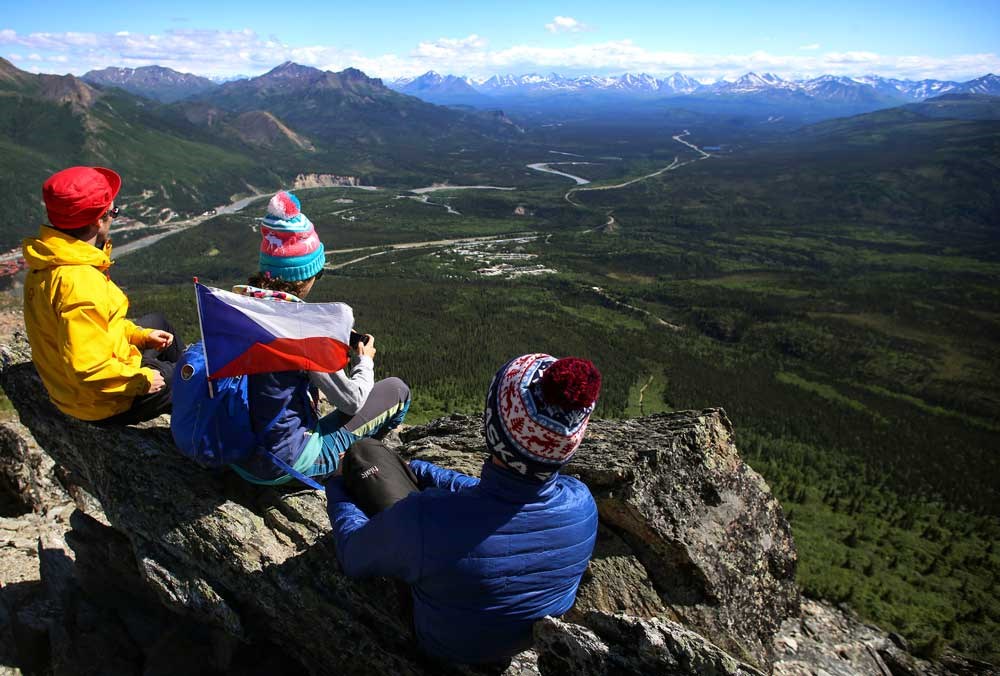 three people sitting on a rocky outcropping, looking out over a landscape of forests, mountains and roads