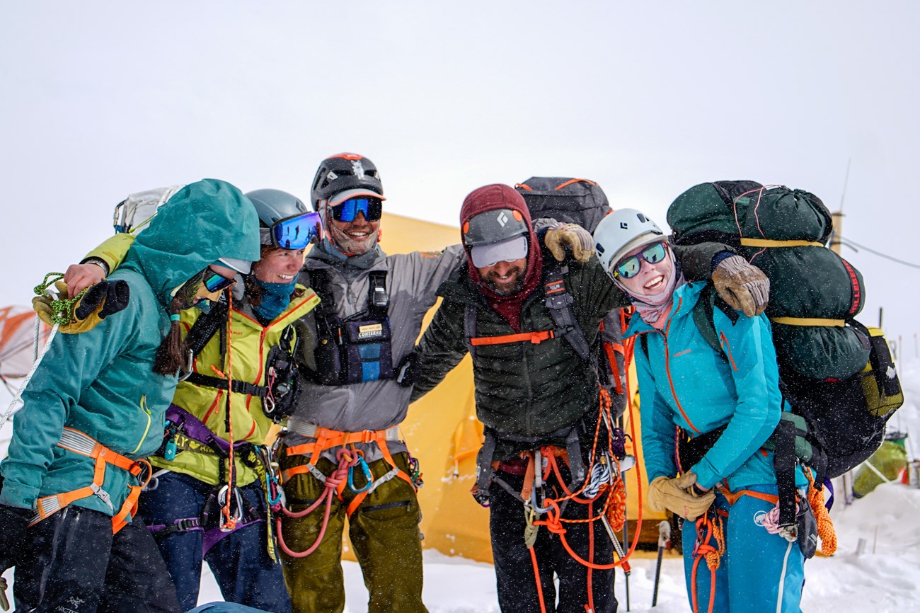 Five teammates in harnesses and climbing gear huddle up in front of a tent