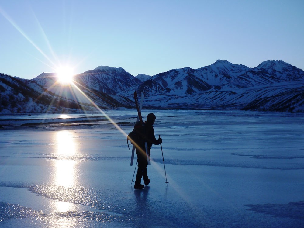 a person with skis strapped on her back walks across a frozen, icy river or lake