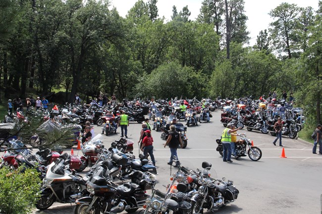 parked motorcycles fill the parking lots with people crowded throughout; volunteers direct motorists where to park