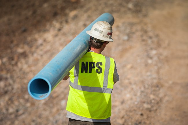 A man wearing a white hardhat and green safety vest with the letters NPS on the back walks away from the camera in a desert setting while carrying a long blue pipe on his shoulder.
