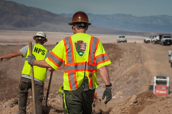 Two workers, both in high-visibility vests and hard hats, stand on rough dirt terrain. The central worker, his back to the camera, wears a vest emblazoned with the National Park Service logo. The expansive, arid landscape and a faraway vehicle.