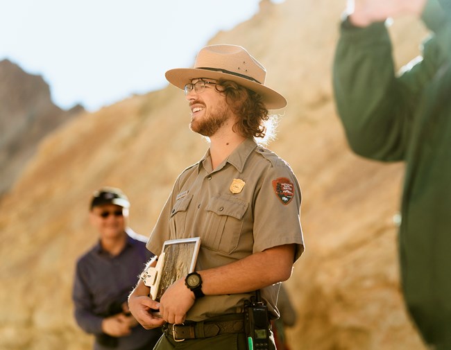 A smiling park ranger stands outdoors in sunlight, holding a folder and clipboard. The background shows another casually dressed person and a blurred natural landscape. The scene gives an impression of an educational talk or tour.
