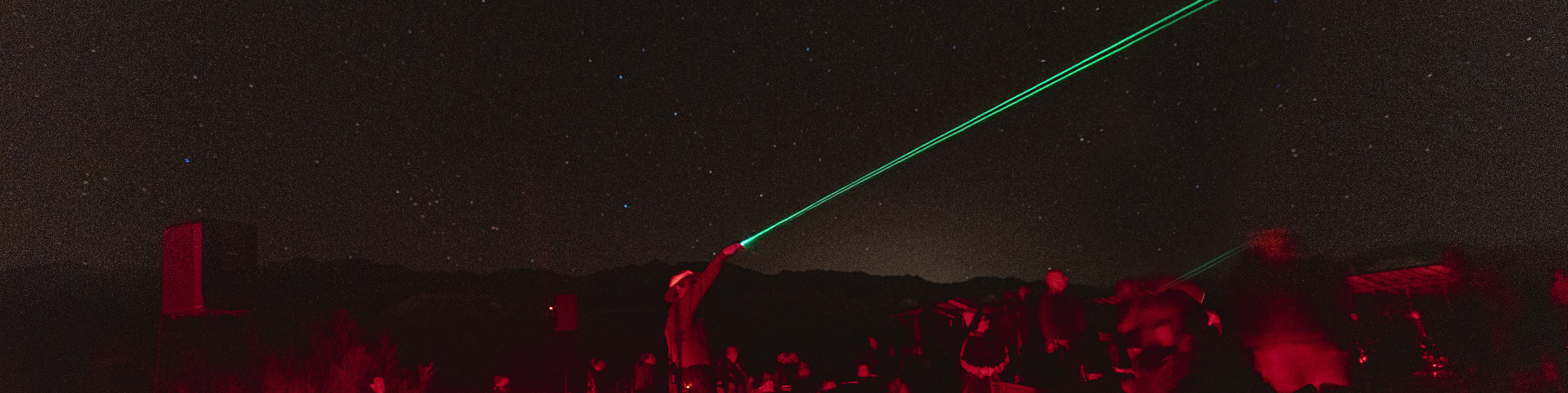 A star-filled sky. A prominent green laser beam stretches diagonally from the bottom-right to the upper-left. Silhouettes of people or objects beneath are faintly lit in red hues.