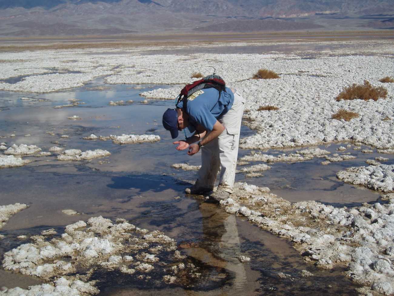 A man standing on white salt bends over a pool of water looking at something in his hand.