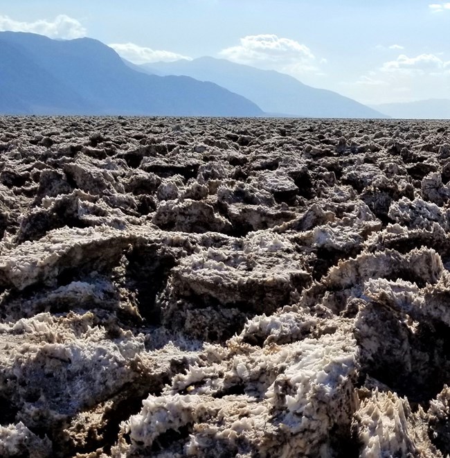 An expansive salt flat with sharp, jagged formations stretches out in the foreground. In the background, mountain ranges rise under a sky with scattered clouds.