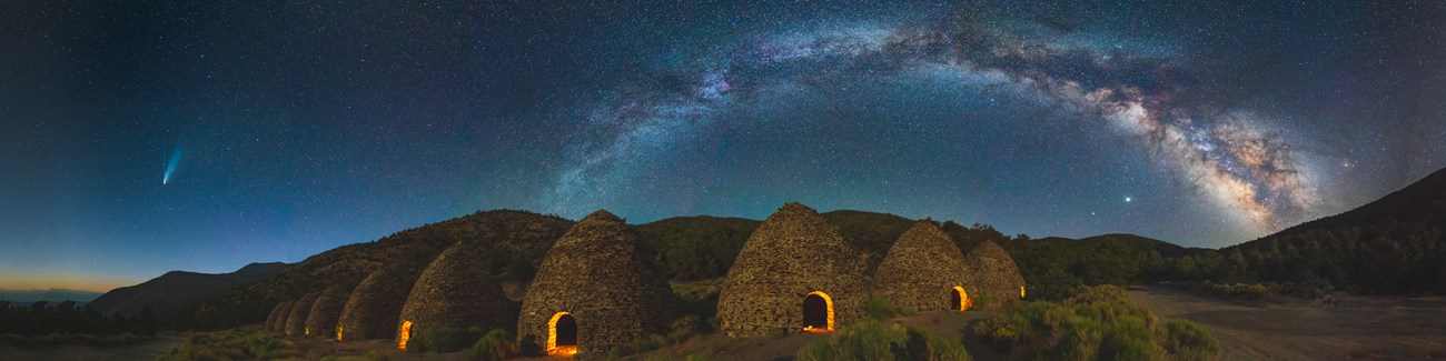 Panorama of ten beehive shaped masonry charcoal kiln structures at night with yellow light shining through the doorways, the milkyway to the right and the Neowise comet to the left.