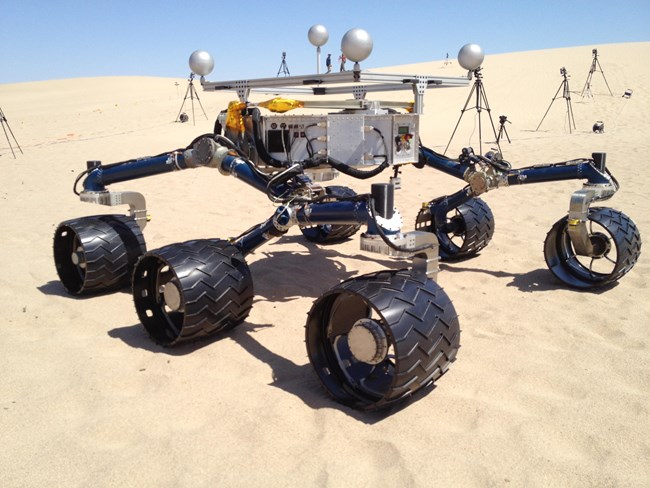 The image displays a six-wheeled robotic vehicle on sand, with scientific equipment in the background. It operates in an expansive desert terrain.