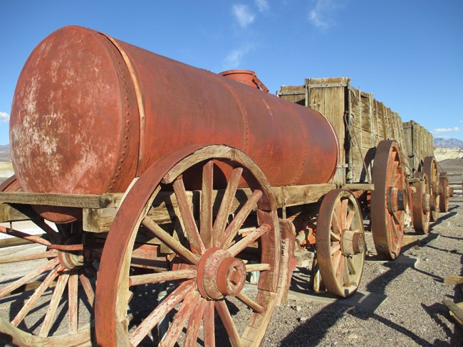 A large rusty tank on large wooden wagon wheels with two large wooden carts behind it.